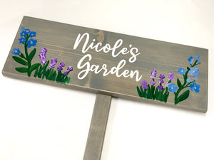 Large custom wooden garden sign painted gray with white cursive letters and hand-painted blue and purple flowers on wood grain background