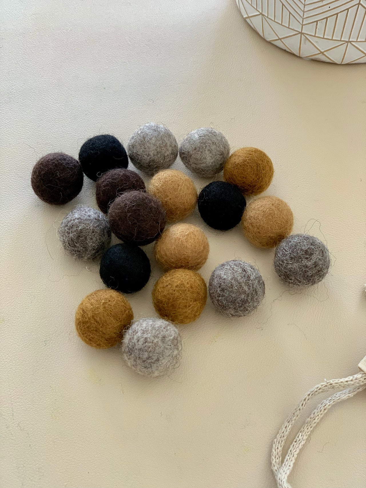 Assortment of 18 felt balls in soil and rock colors on light surface with partial view of patterned container and string rope