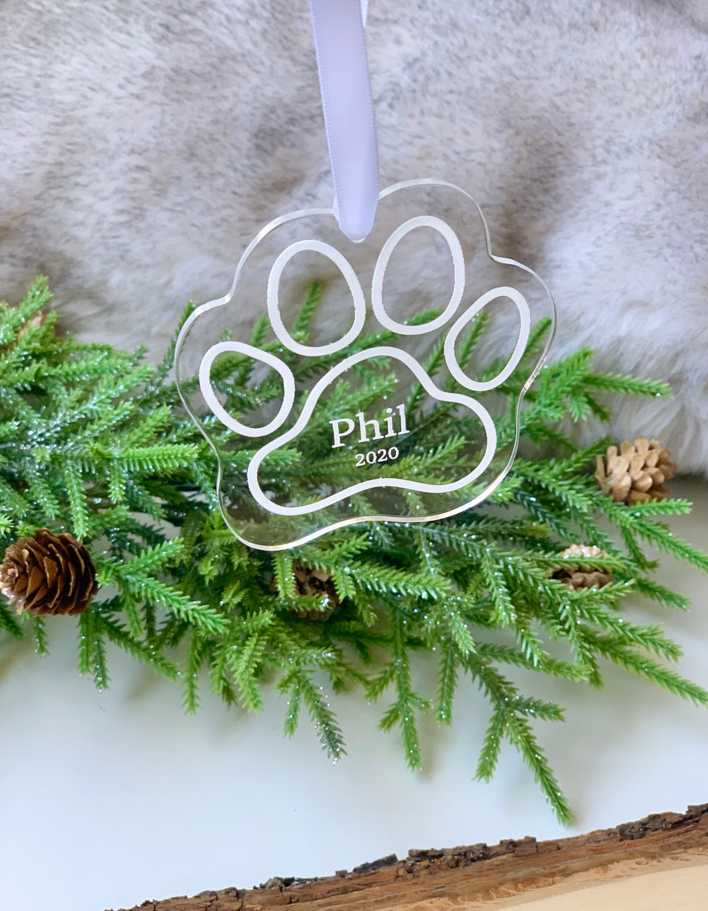 Clear acrylic paw print Christmas ornament engraved with name Phil and year 2020 hanging on white ribbon above green pine branches