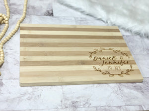 Rectangular striped wooden cutting board with personalized engraved names and date inside wreath design on bottom right corner