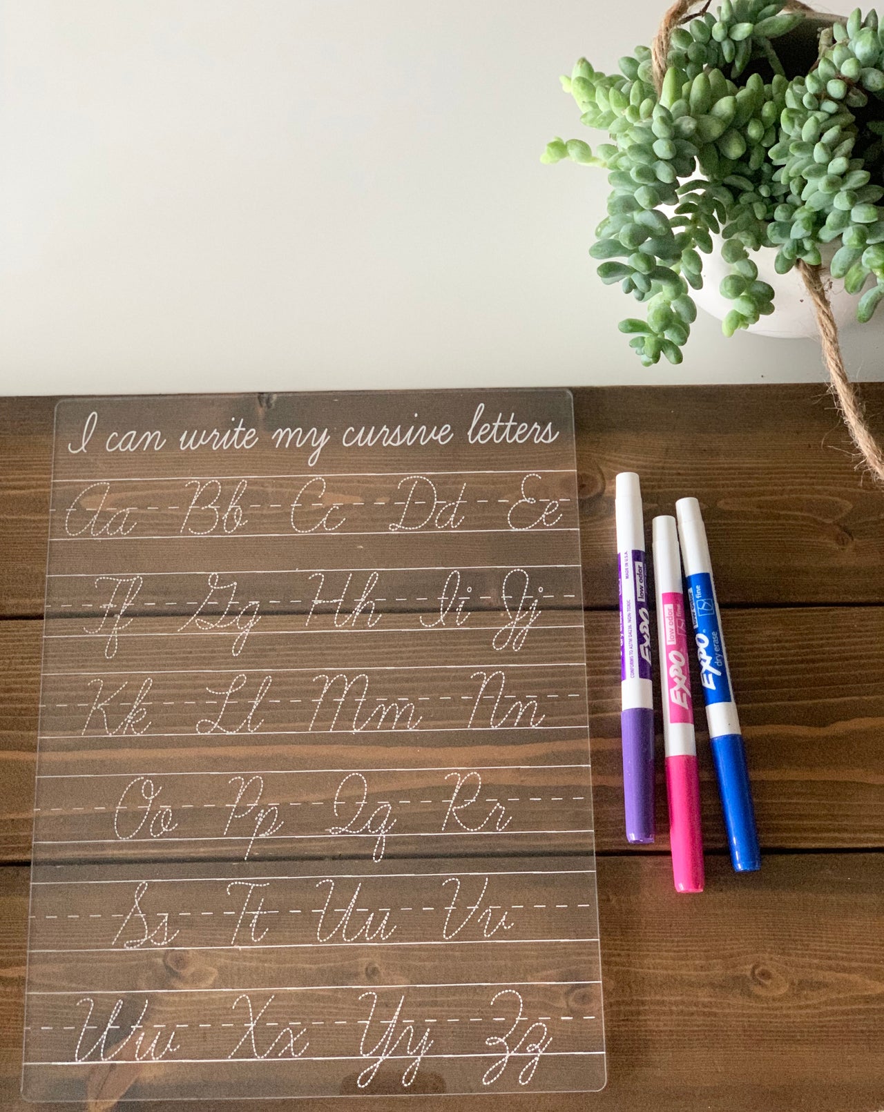 Transparent acrylic dry erase board engraved with cursive alphabet letters and three colored markers on wooden surface