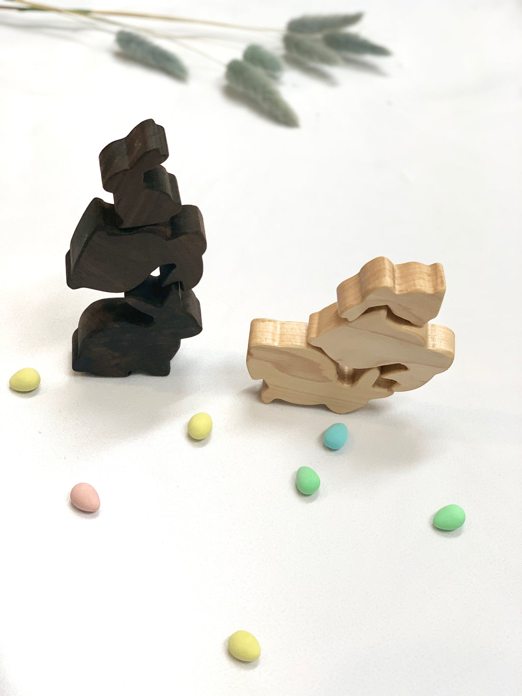 Stacked wooden toy bunnies in natural and dark wood finishes with scattered pastel-colored candy eggs on white surface
