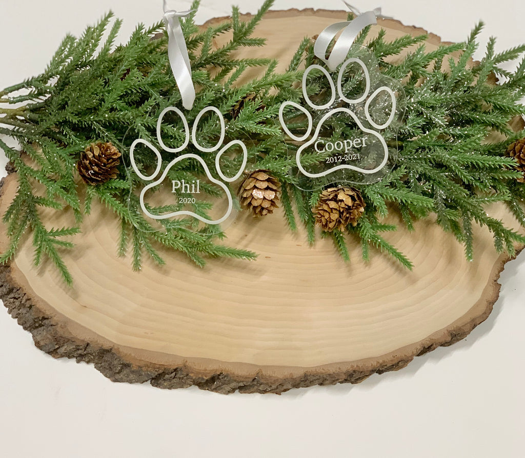 Clear acrylic paw print pet memorial ornaments engraved with names and dates hanging on greenery with pinecones