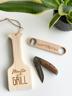 Custom wooden grill spatula with "Master of the Grill" text, personalized wooden bottle opener, and folding pocket knife on white surface