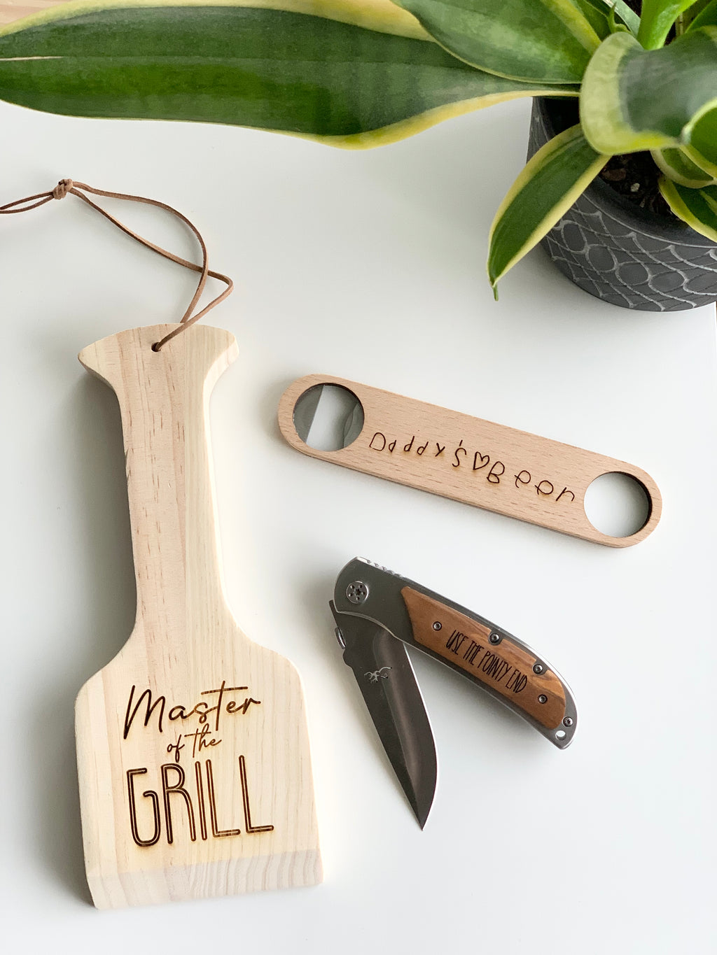 Custom wooden grill spatula with "Master of the Grill" text, personalized wooden bottle opener, and folding pocket knife on white surface