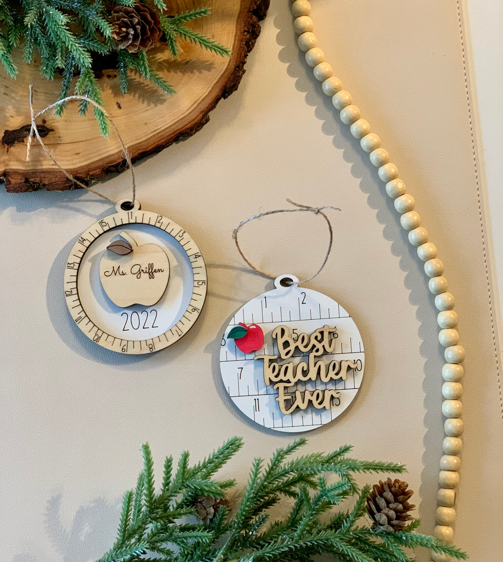 Two round wooden Christmas ornaments with ruler design, one with red apple and "Best teacher ever" text, the other engraved with apple and name