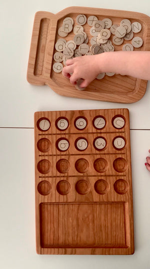 Natural wooden 20 frame sensory tray with engraved alphabet discs scattered on matching wooden tray held by child’s hand on white surface