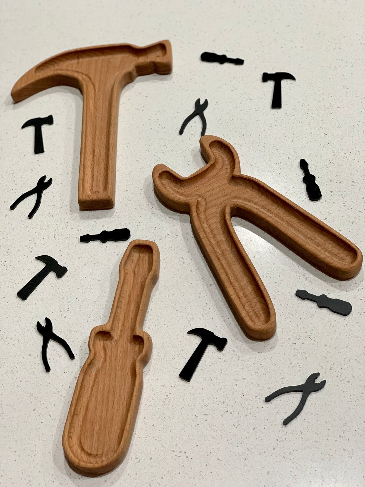 Wooden tool-shaped trays with black acrylic loose parts in hammer, wrench, screwdriver, and plier shapes on white surface