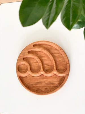 Round wooden sensory tray carved with wave shapes on white surface with green leaves overhead