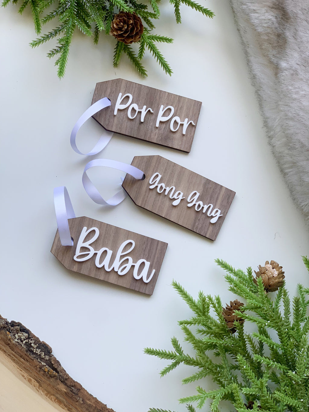 Set of three custom wooden gift tags with white cursive lettering and white ribbons on white surface with pine accents