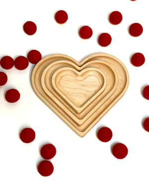 Natural wooden nesting heart plates stacked inside each other surrounded by scattered red pom-poms on white background