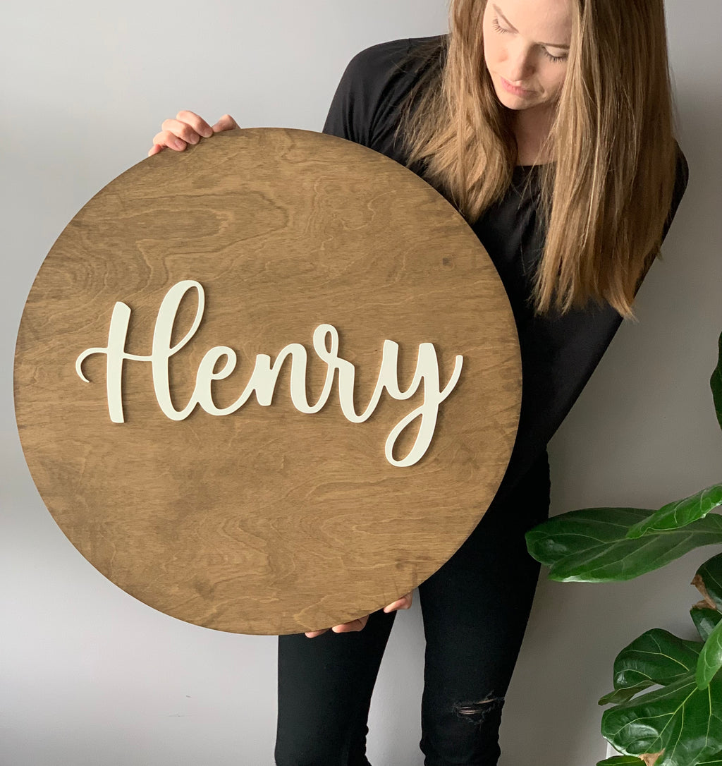 Round wooden name sign with white cursive letters spelling Henry held by woman in black outfit