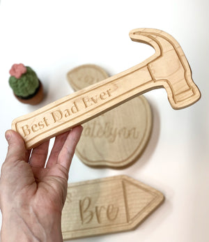 Light natural wooden hammer-shaped plaque engraved with "Best Dad Ever" held above engraved wooden name signs on white background