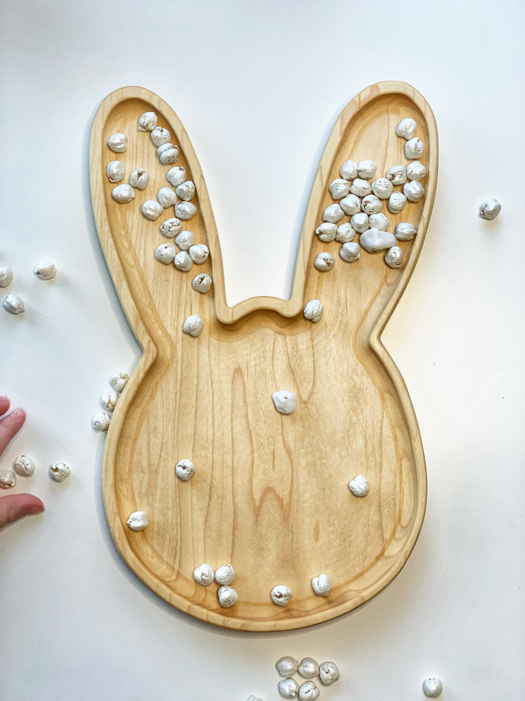 Large natural wooden bunny-shaped plate with scattered white popcorn kernels on white surface and child’s hand nearby
