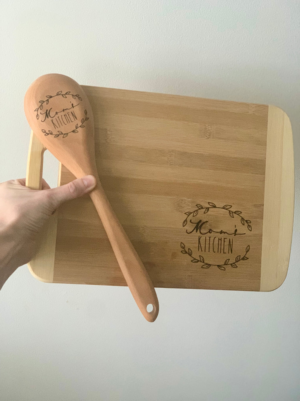 Personalized bamboo cutting board and wooden spoon set engraved with "Mom's Kitchen" and decorative leaf wreath designs