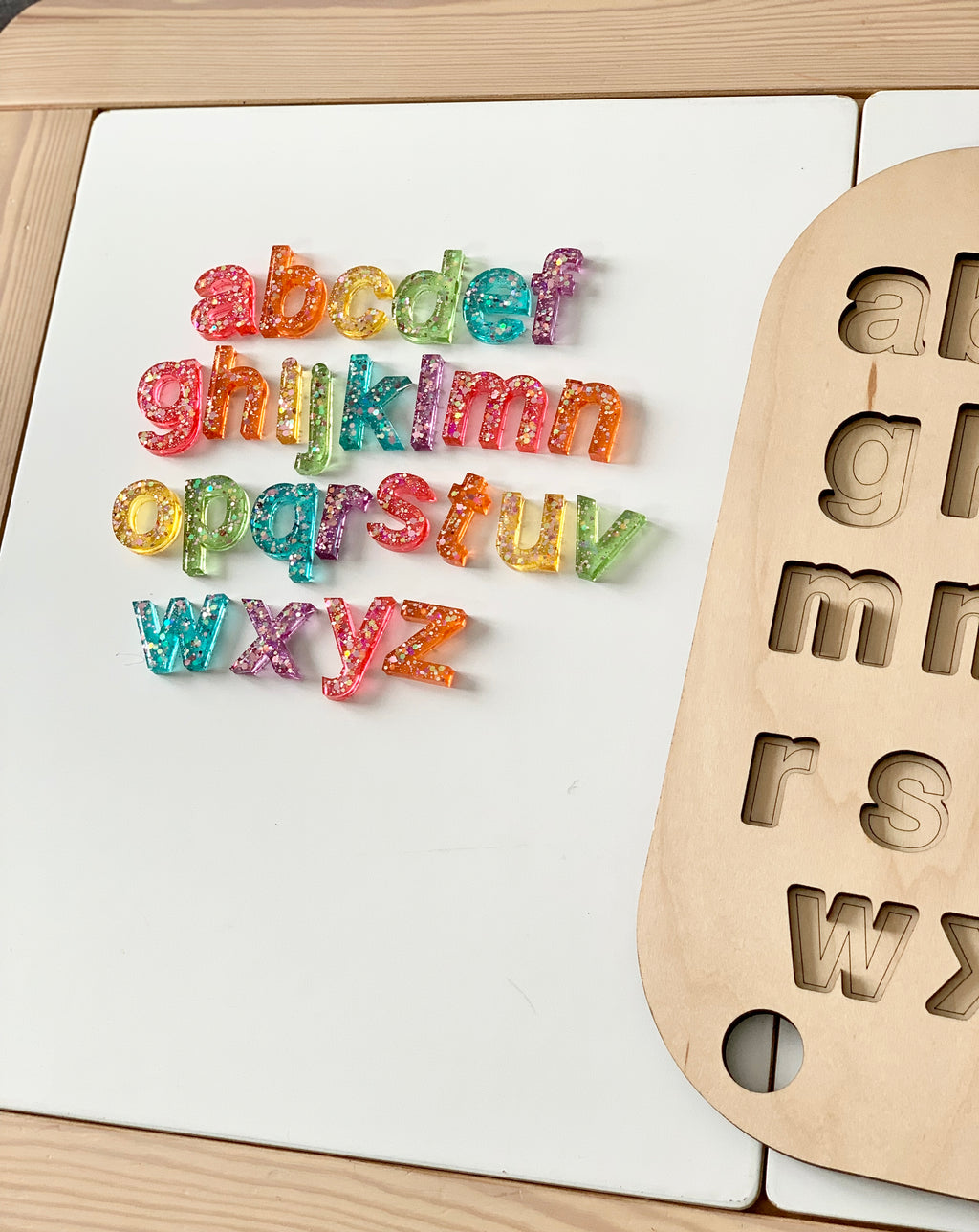 Lowercase resin alphabet letters in multicolor glitter beside a wooden Flisat puzzle board with matching letter cutouts on white table