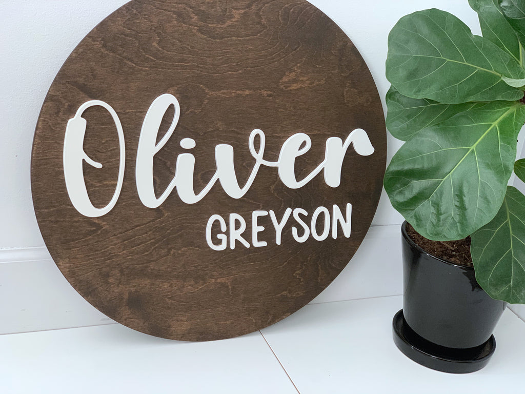 Round wooden personalized name sign with white cursive and block letters on dark stained wood background next to green plant
