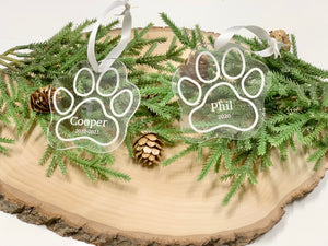 Clear acrylic paw print pet memorial Christmas ornaments engraved with names and dates on wooden slab with greenery