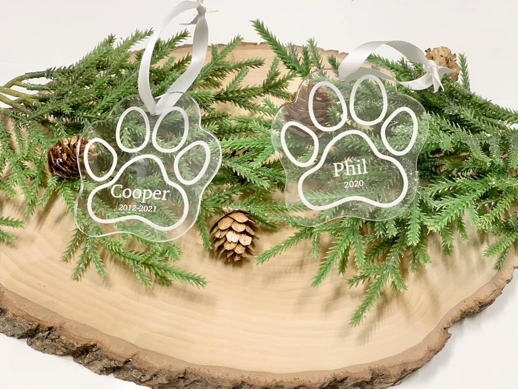 Clear acrylic paw print pet memorial Christmas ornaments engraved with names and dates on wooden slab with greenery