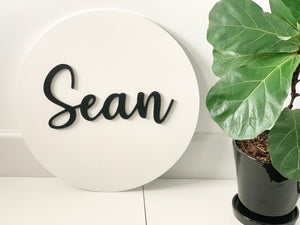 3D round wooden name sign painted white with black cursive letters spelling Sean beside green potted plant