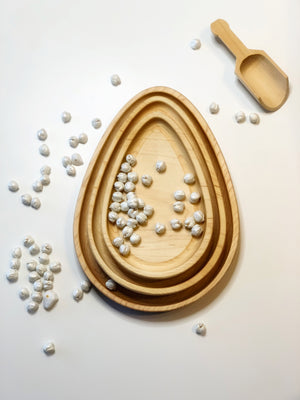 Set of natural wooden nesting egg plates with small white sensory beads and wooden scoop on white surface
