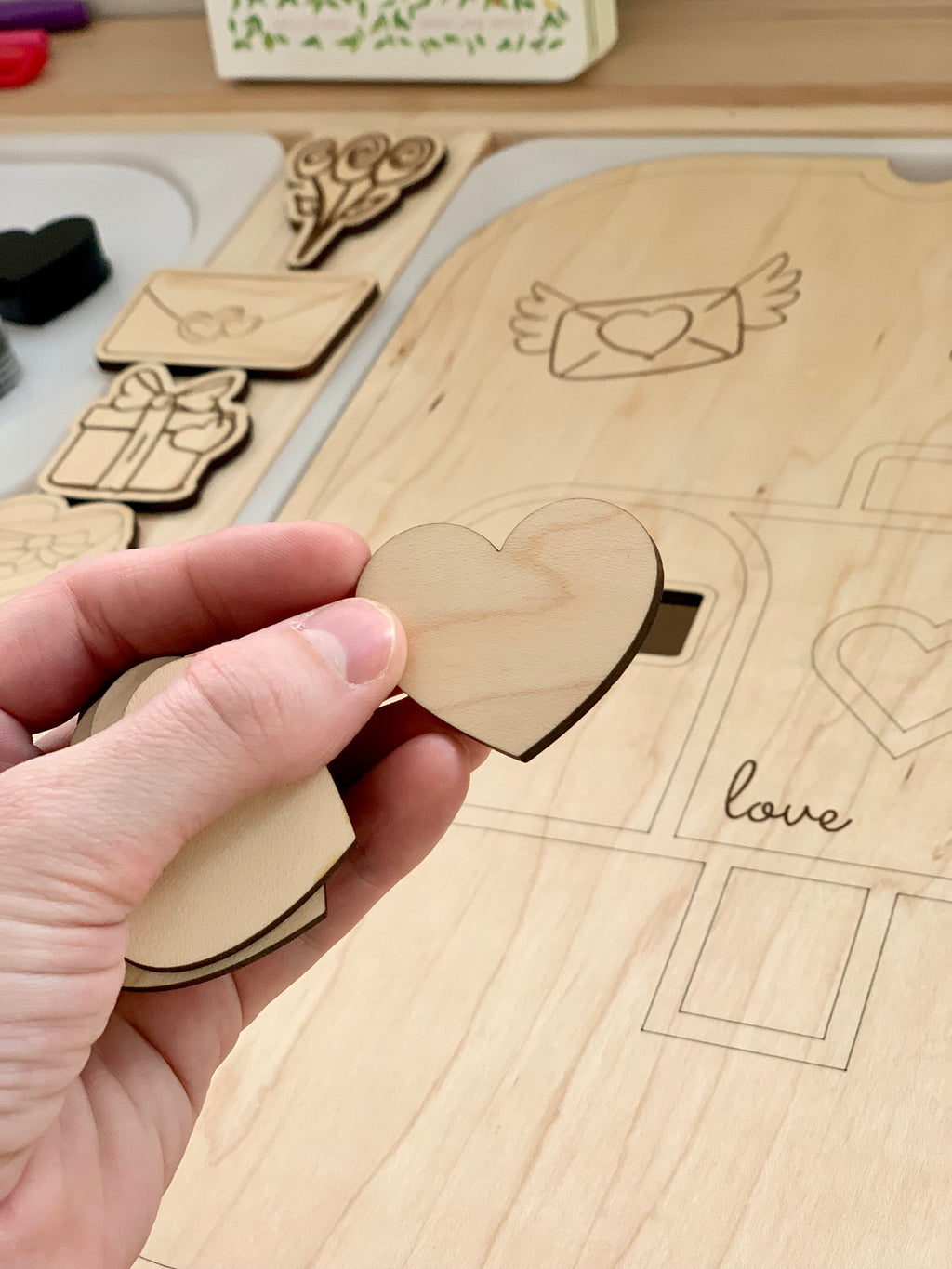 Hand holding wooden heart-shaped laser cut piece over matching puzzle board with love-themed shapes on light wood surface