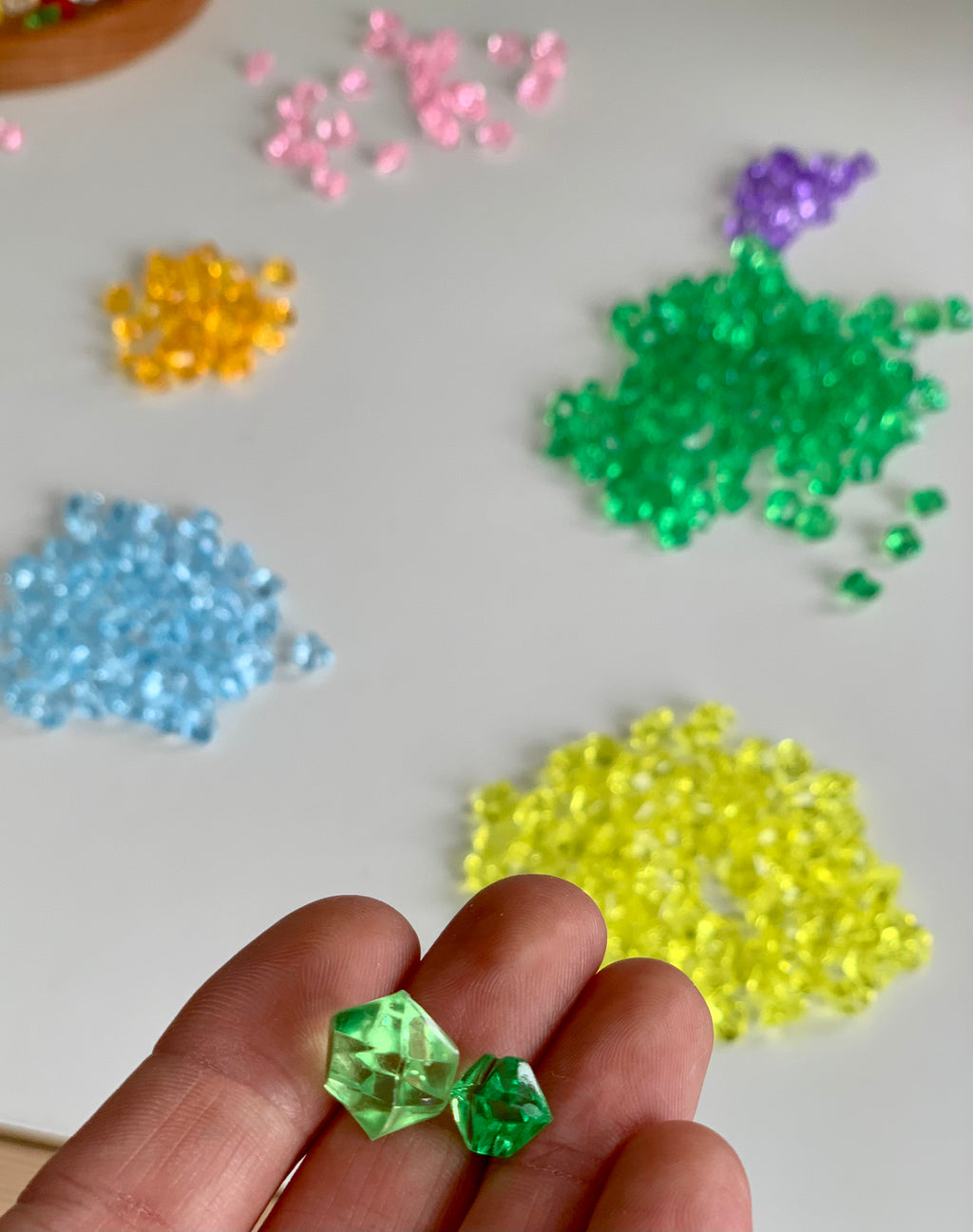 Assorted colorful acrylic treasure gems in various sizes held in fingers with scattered piles on white surface