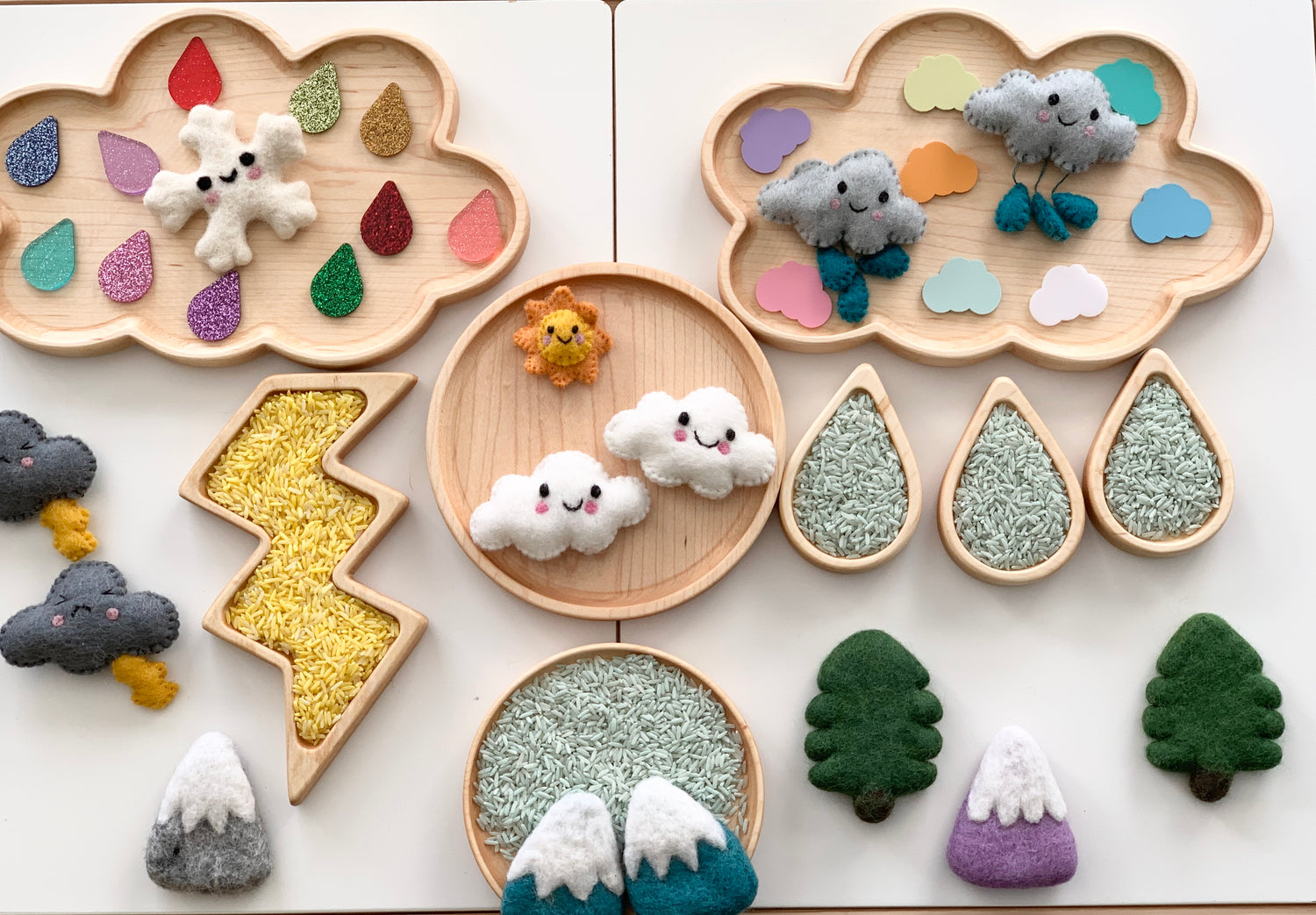 Wooden cloud and round sensory trays with colorful felt raindrops, clouds, trees, lightning bolt, and mountain shapes on white surface