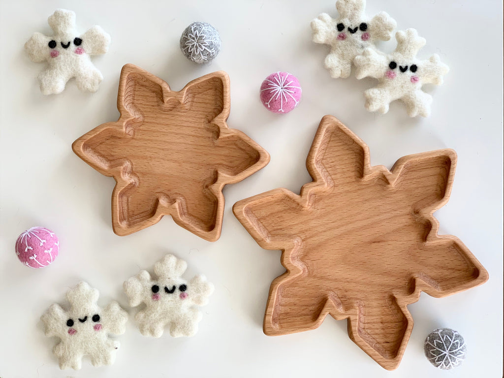 Two natural wooden snowflake-shaped small plates on white surface surrounded by white felt snowman and pink and gray felt balls