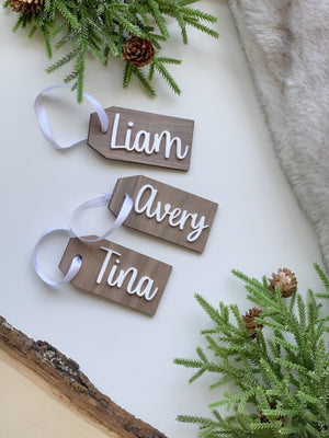 Custom wooden gift tags with white cursive names Liam, Avery, and Tina on natural wood with white ribbons and greenery background