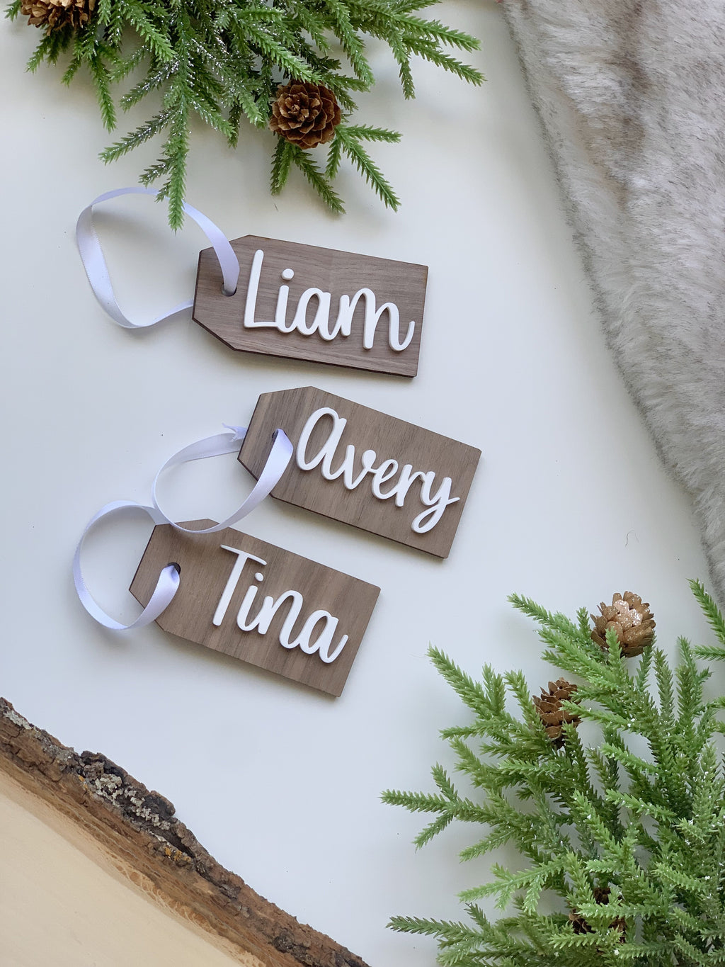 Custom wooden gift tags with white cursive names Liam, Avery, and Tina on natural wood with white ribbons and greenery background