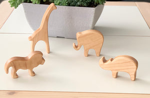 Natural wooden safari animal toy figurines including giraffe, elephant, lion, and rhinoceros on white surface with planter background