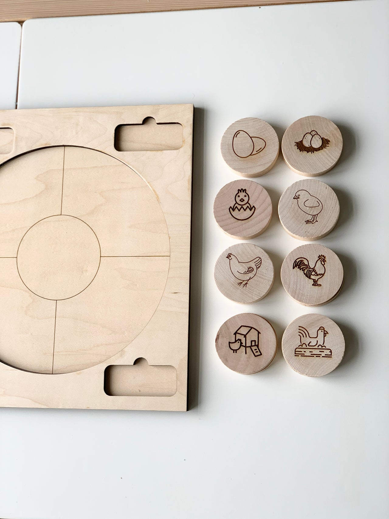 Round natural wooden discs engraved with chicken life cycle stages next to wooden game board with circular sections