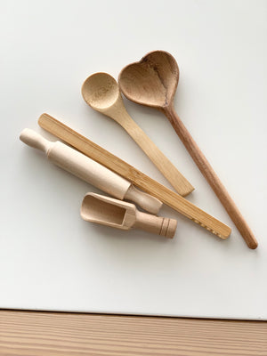 Set of natural wooden sensory bin tools including heart-shaped spoon, round spoon, scoop, rolling pin, and stick on white background